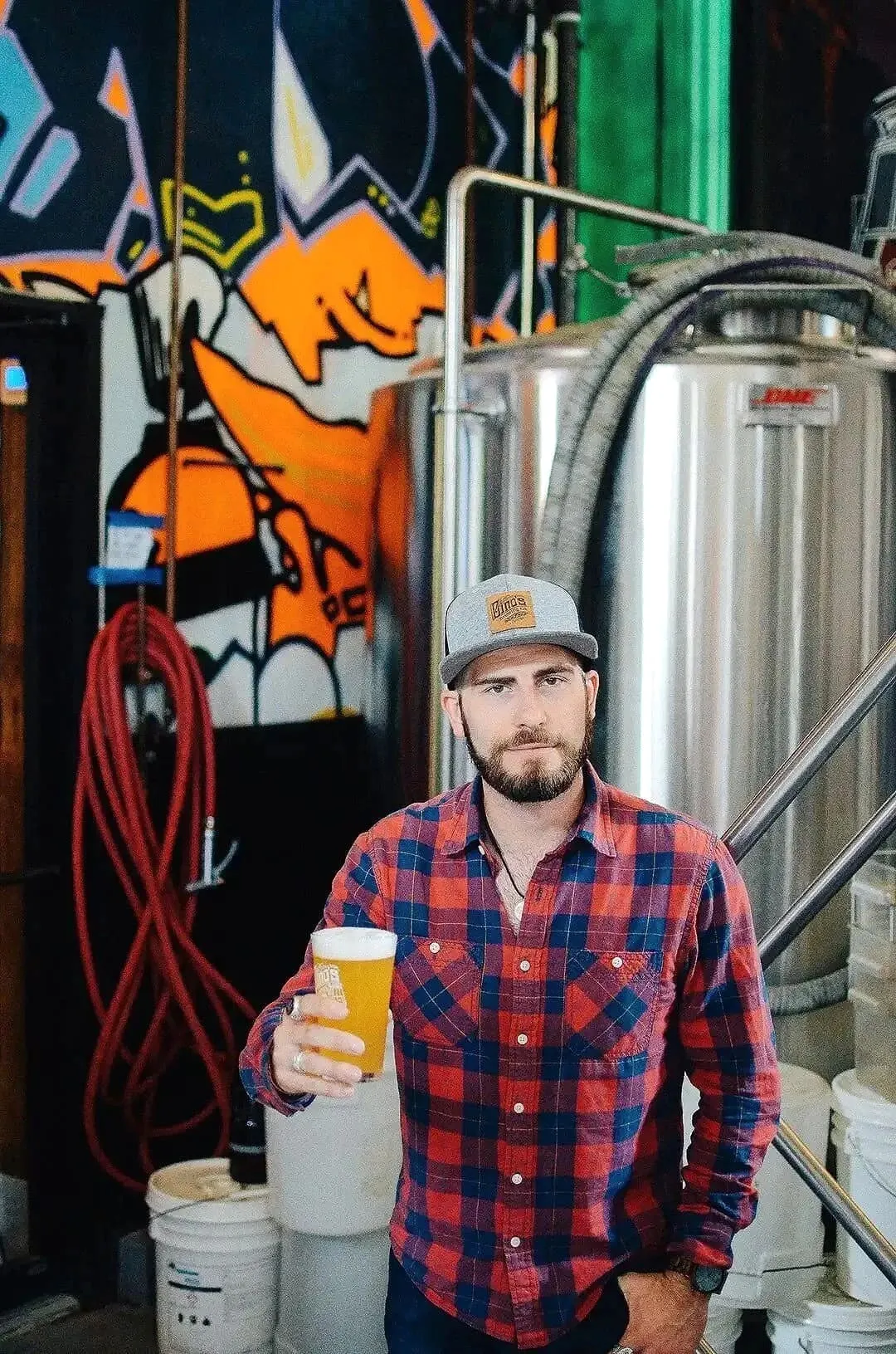 Man holding a glass of beer inside a brewery with fermentation tanks in the background