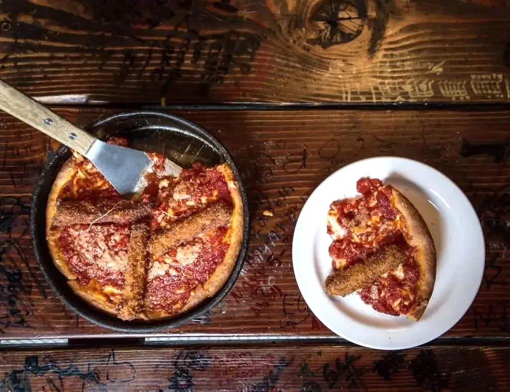 Deep-dish pepperoni pizza slice on white plate beside full cast iron pan pizza on wood table