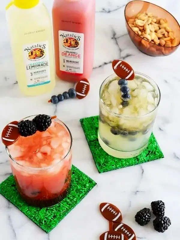 Cocktails with berries and ice on green coasters with lemonade and orange juice bottles in the background