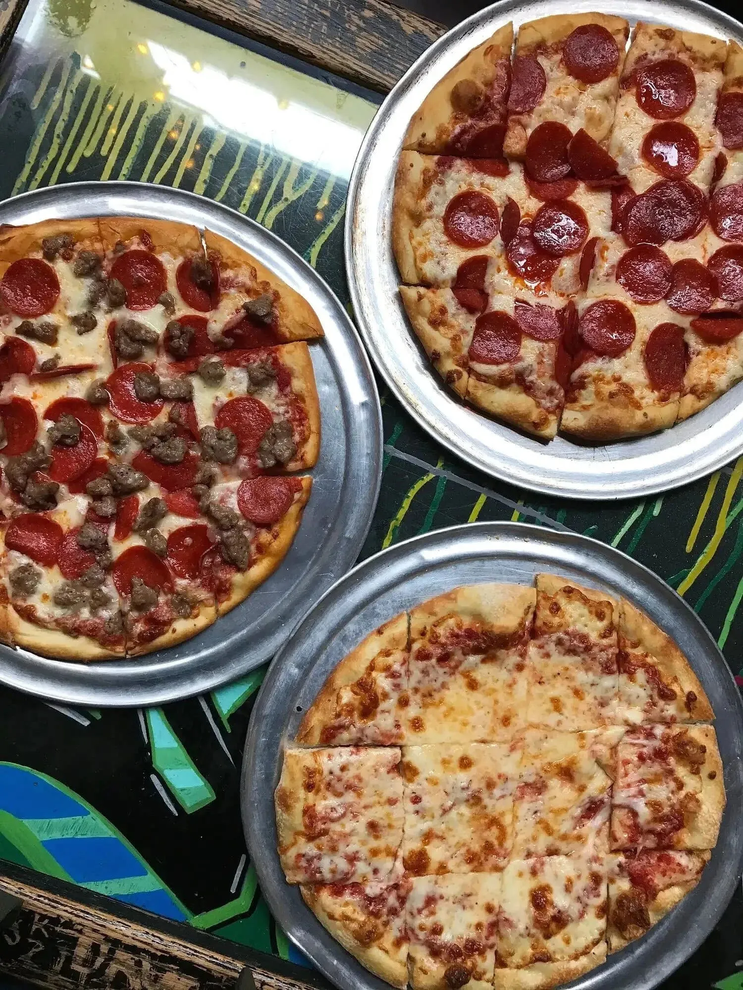 Three tavern-style pizzas cut into squares on metal pans, topped with pepperoni, sausage, and cheese