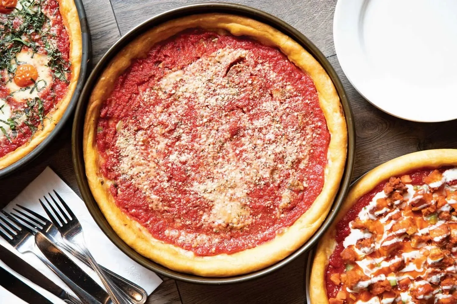 Overhead shot of a deep dish pizza in its pan alongside other pizzas on the table