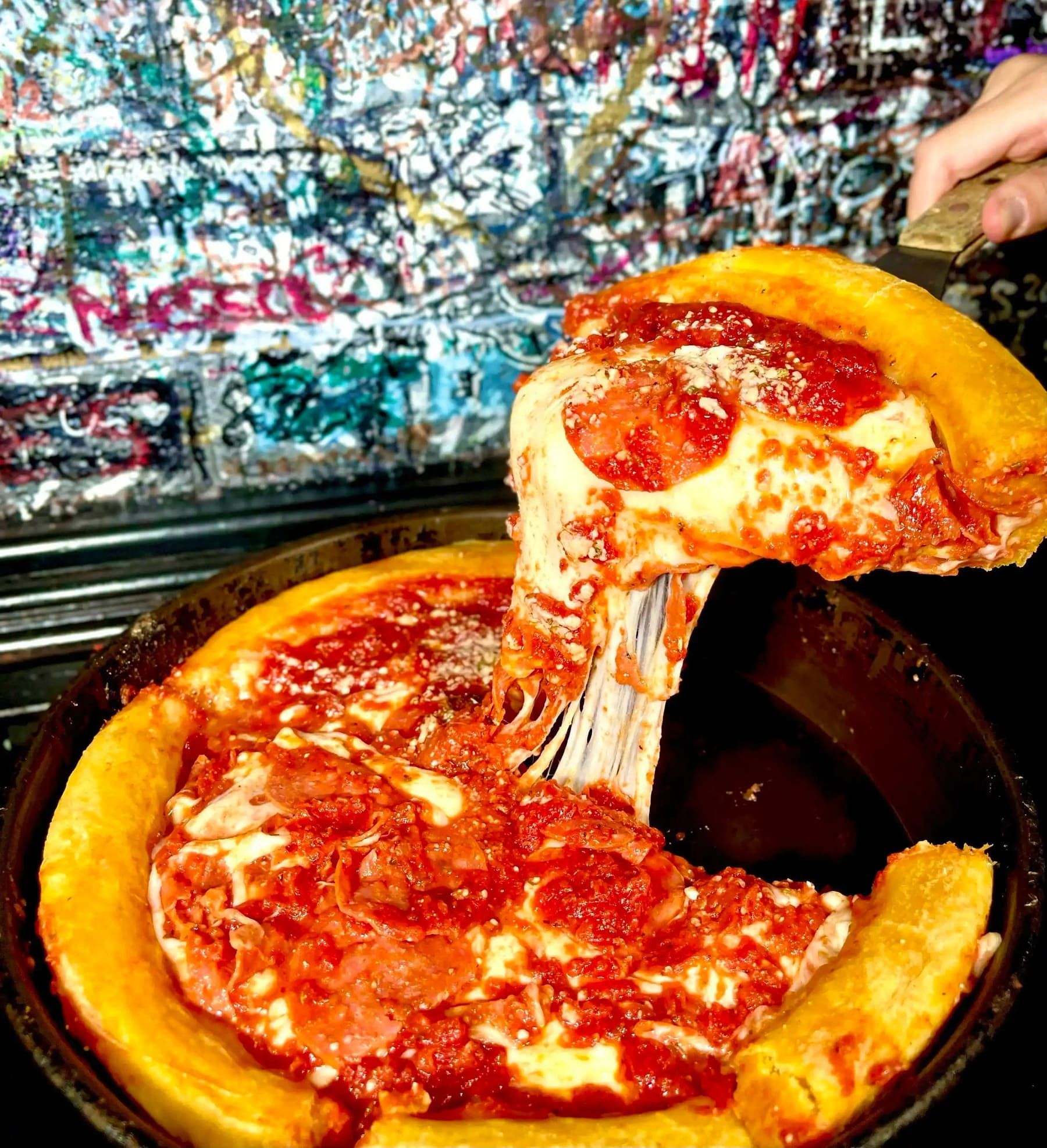 A slice of pepperoni deep dish pizza lifted with a long cheese pull, in front of a graffiti wall
