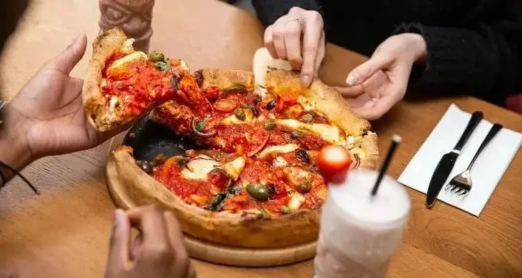 Diners pulling apart a deep dish pizza topped with olives and tomatoes at a wooden table