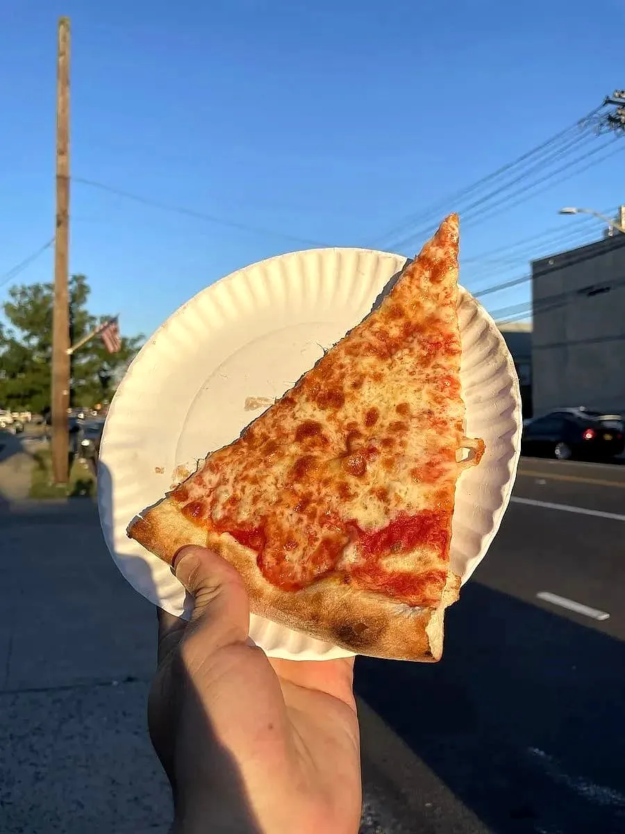 A hand holding a wide slice of New York-style cheese pizza on a paper plate outdoors