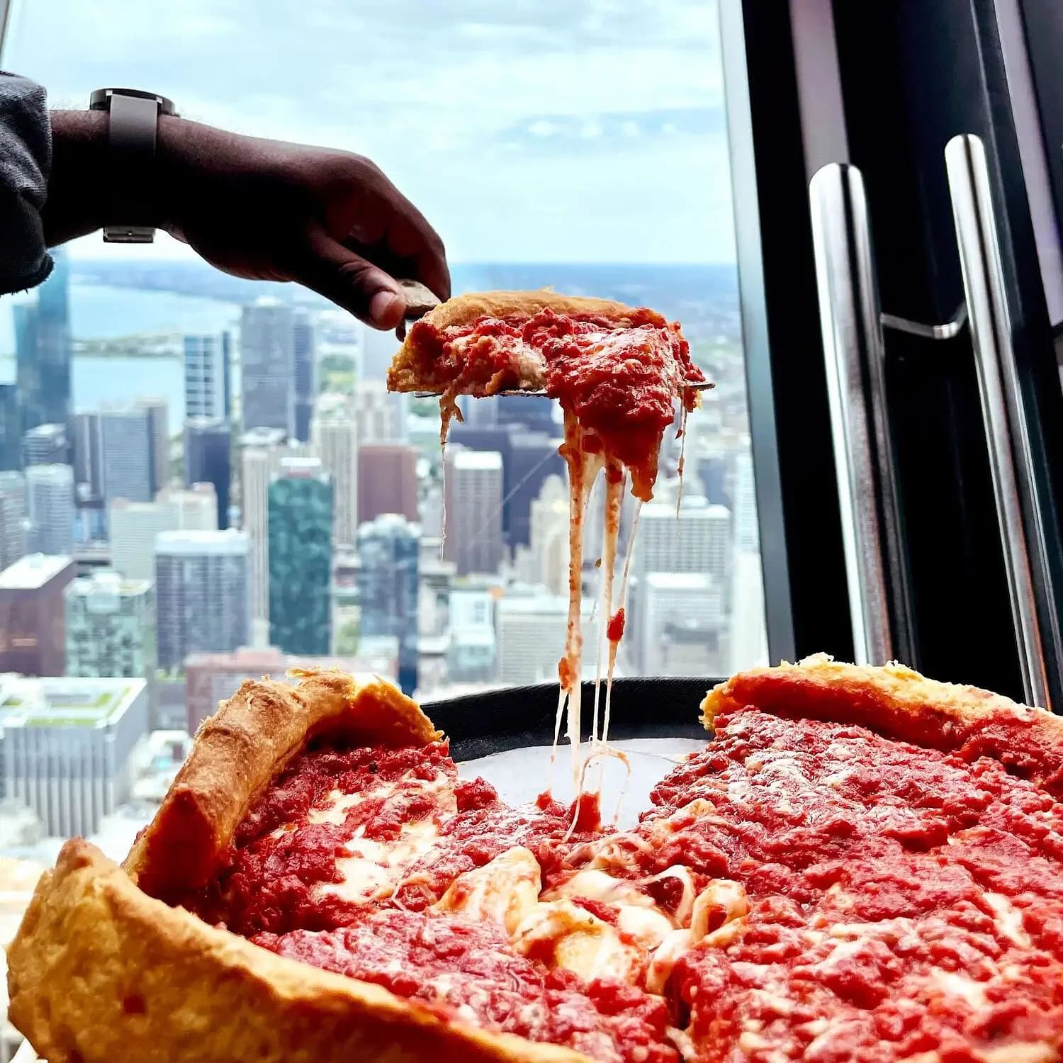 A hand lifting a cheesy slice of deep dish pizza with the Chicago skyline visible through a window at 360 Chicago