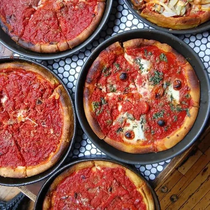 People preparing a pizza with various toppings including green peppers, mushrooms, onions, cooked sausage, pepperoni, and cheese, with multiple bowls of sauce and dough.