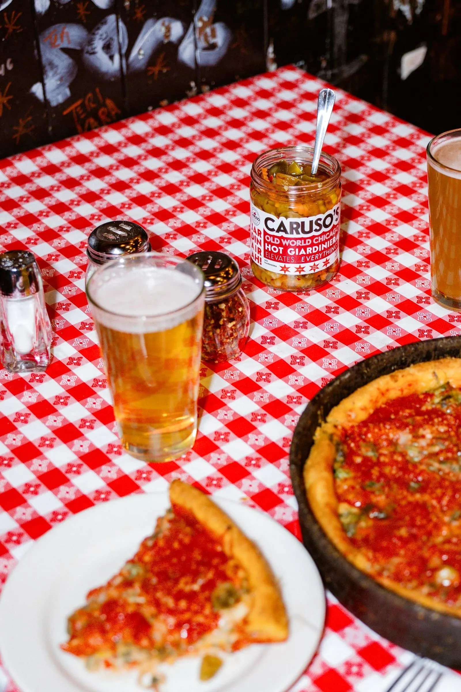 A jar of Caruso’s giardiniera next to a beer and a slice of deep dish pizza on a checkered tablecloth