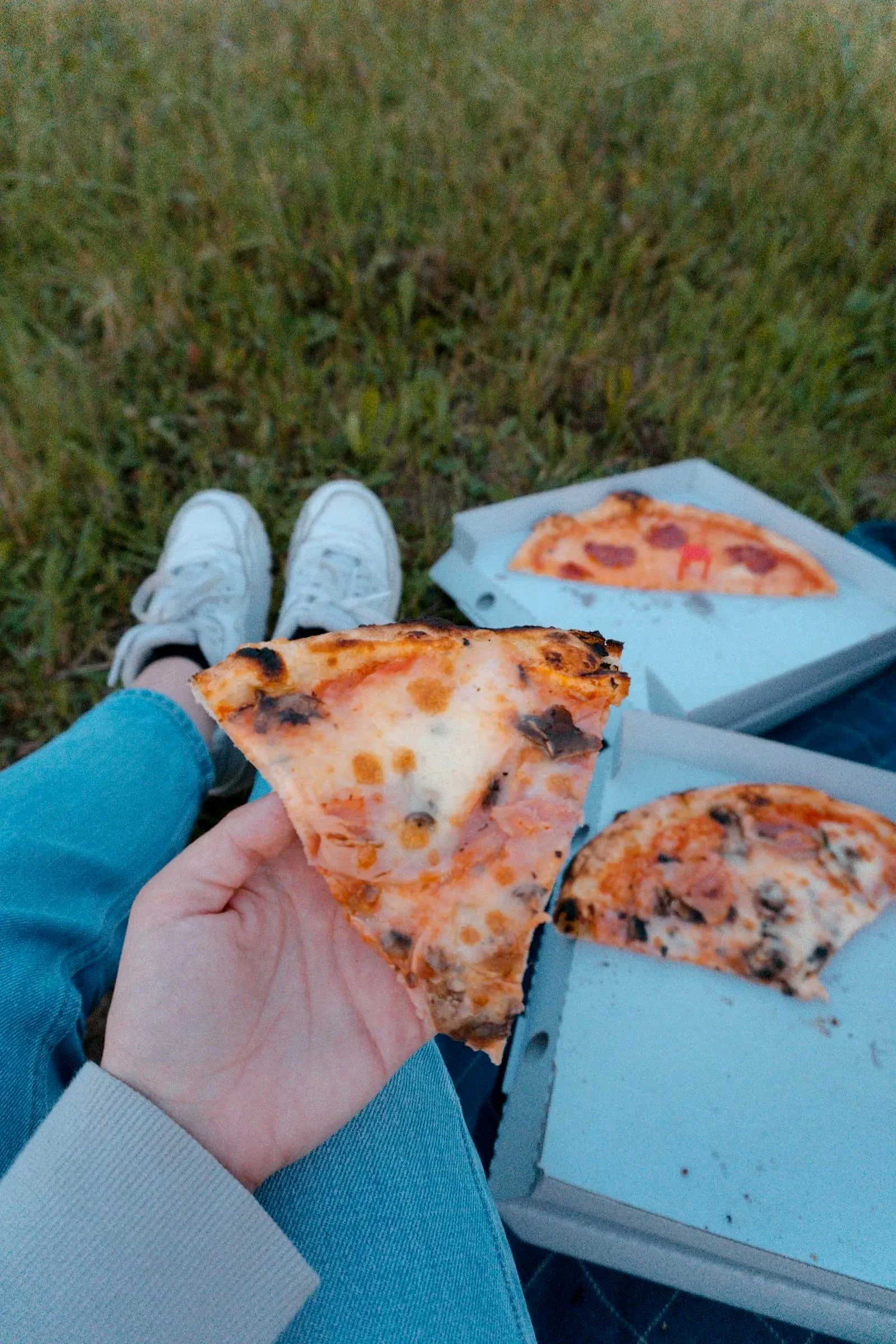 A person sitting outdoors on grass holding a folded slice of pizza next to open pizza boxes