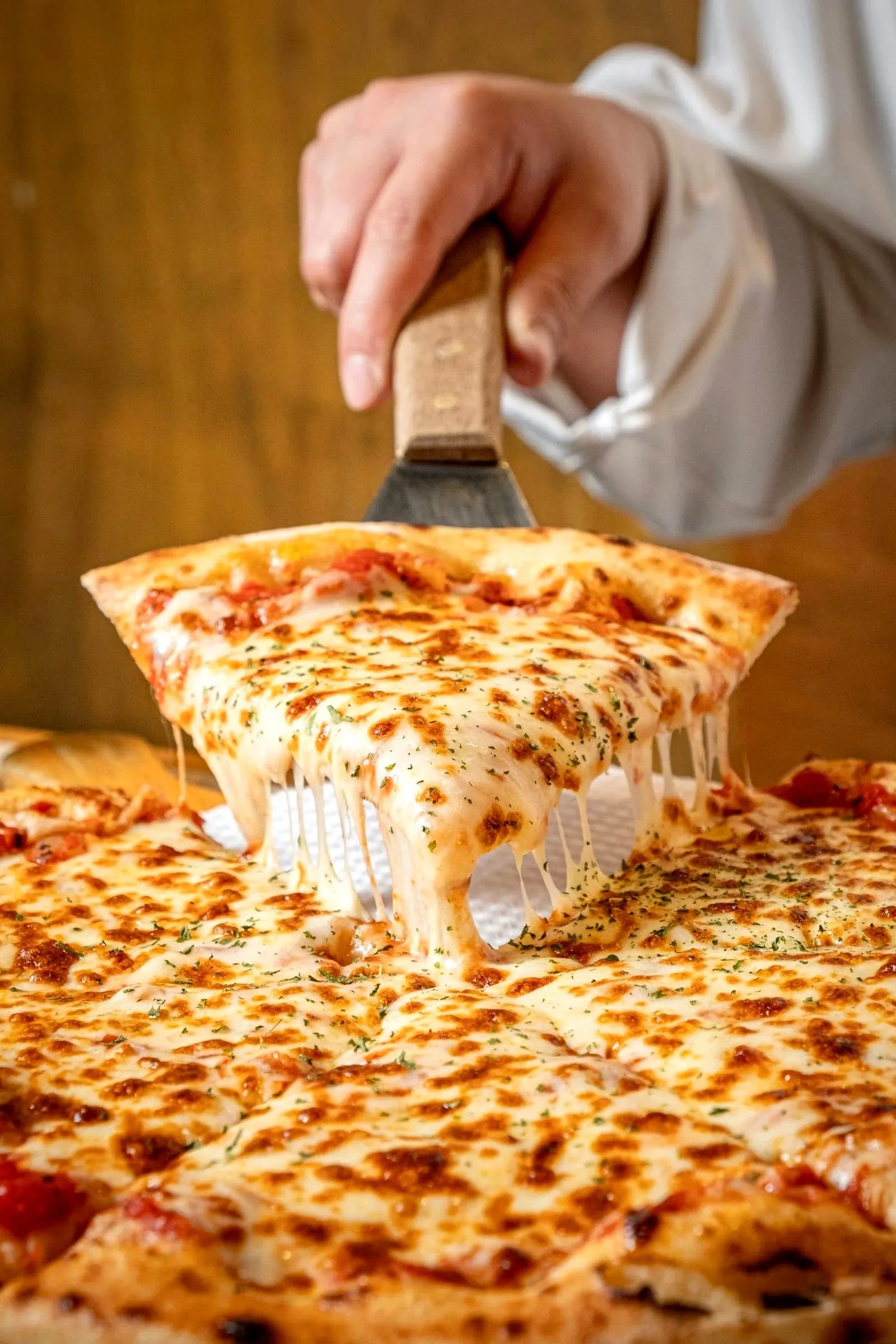 A chef lifting a stretchy cheese slice from a large sheet pizza with a metal spatula
