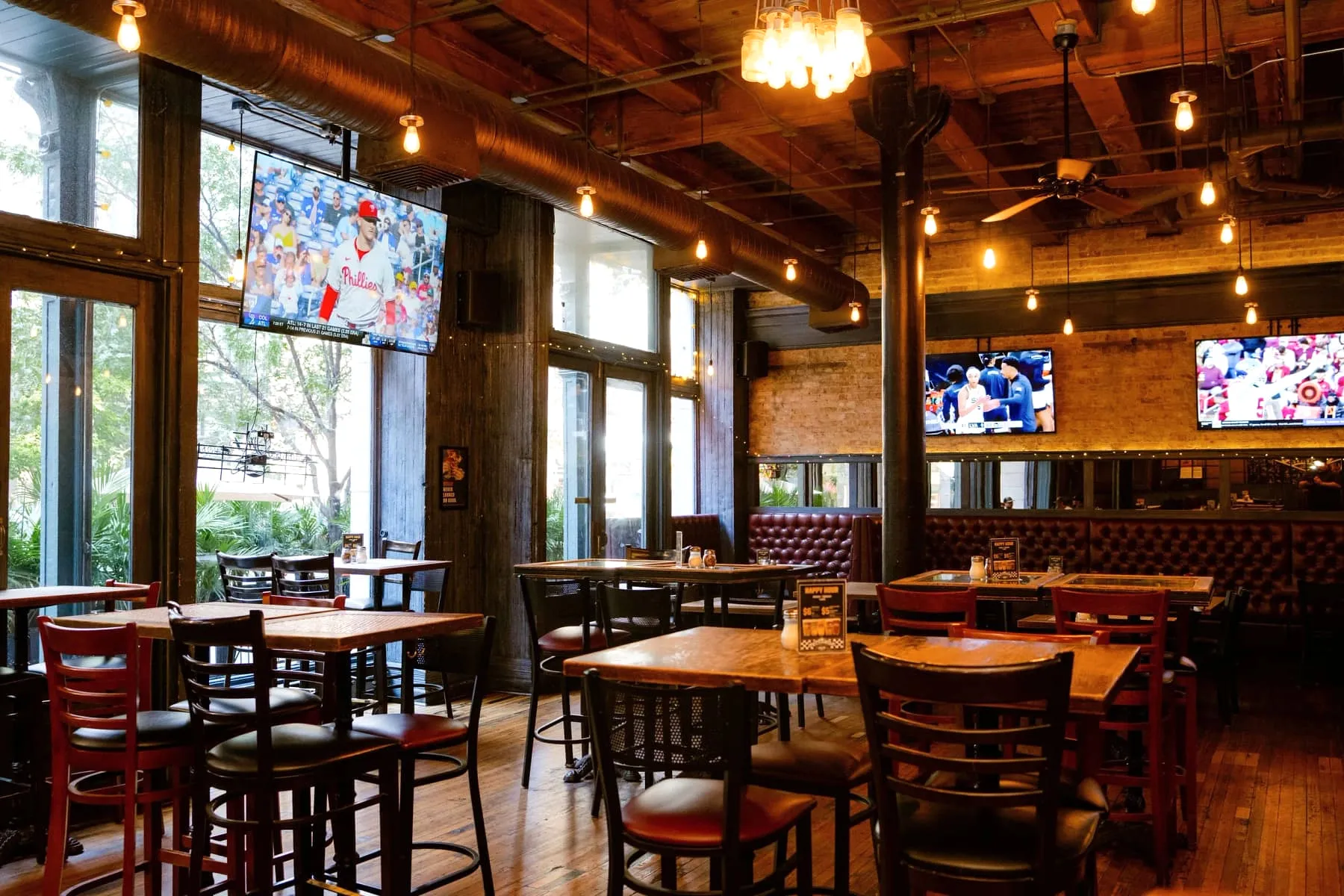 Sunlit dining room at Gino’s East South Loop with exposed brick, timber ceiling beams, Edison bulbs, tufted booths and baseball on the TVs