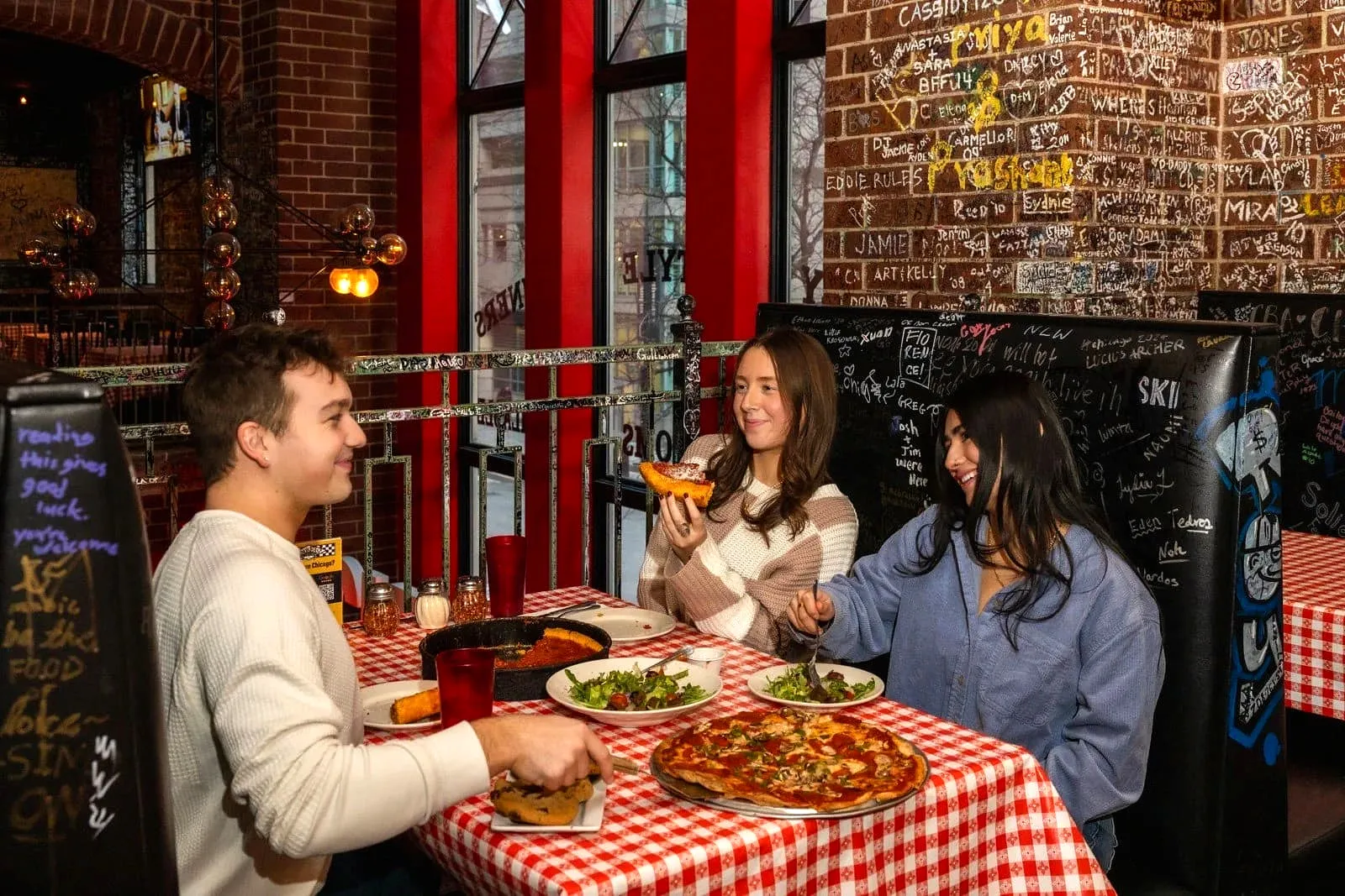 Three friends sharing deep dish pizza and salads at a booth in front of a graffiti-covered brick wall
