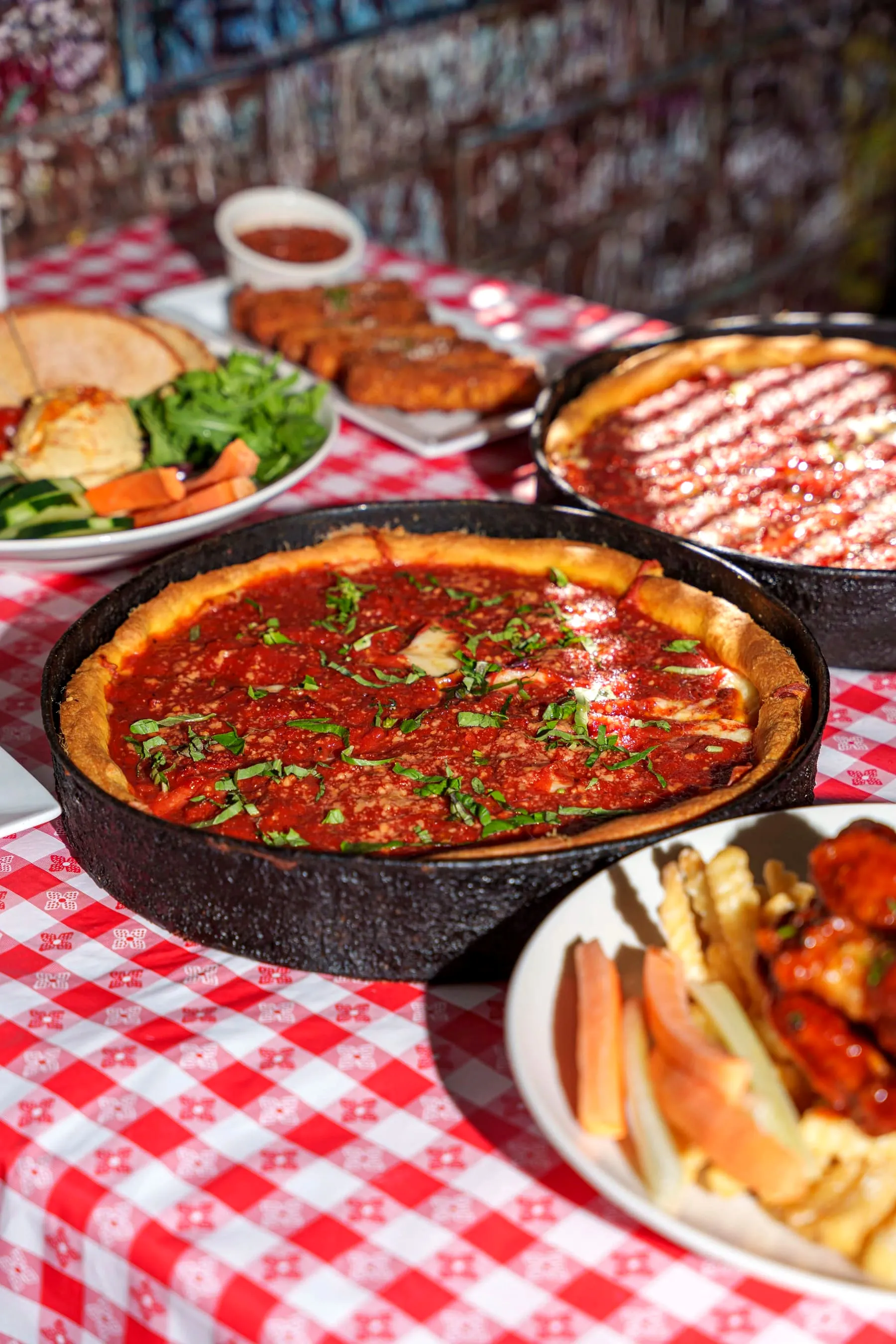 Two Gino’s East deep dish pizzas in black pans on a red-checkered tablecloth, with salads and appetizers and a graffiti wall behind