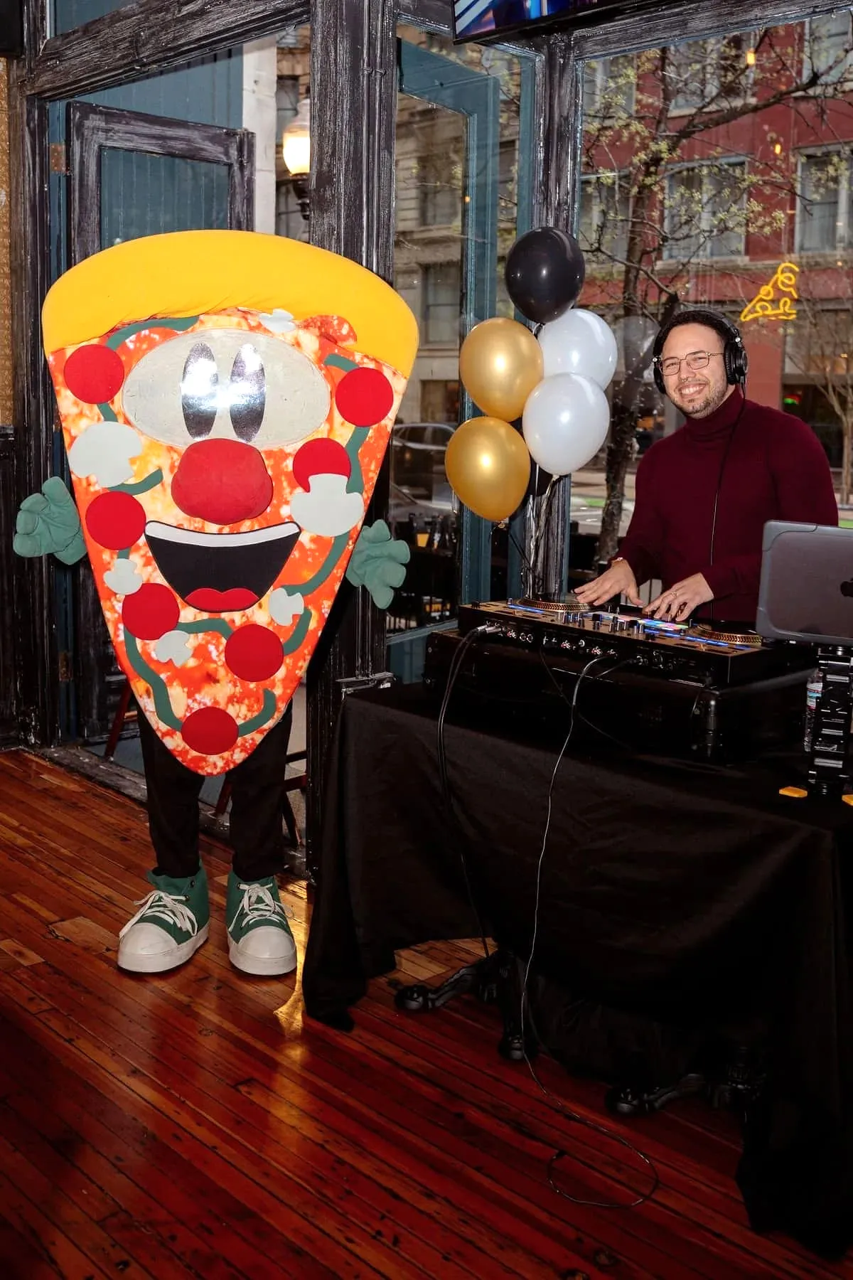 A giant pizza slice mascot posing beside a DJ spinning music at the South Loop reopening party