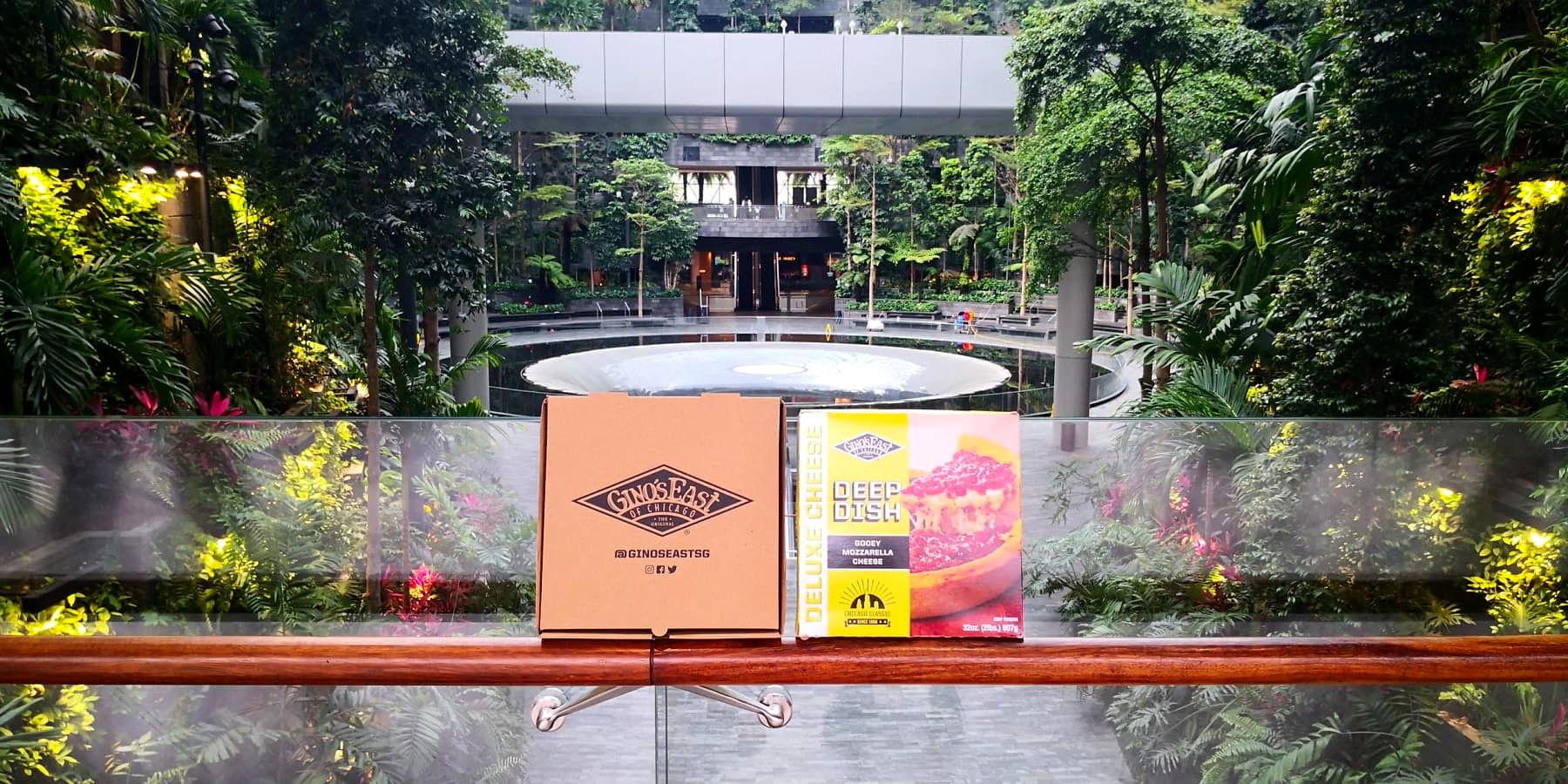 A Gino's East pizza box and frozen deep dish cheese pizza box displayed on a railing overlooking the Jewel Changi Airport forest valley