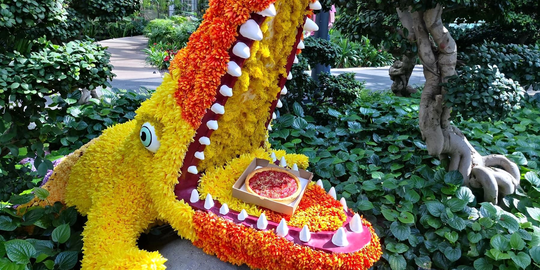 A whimsical flower dragon sculpture holding a Gino's East deep dish pizza box in its mouth at a Singapore garden display
