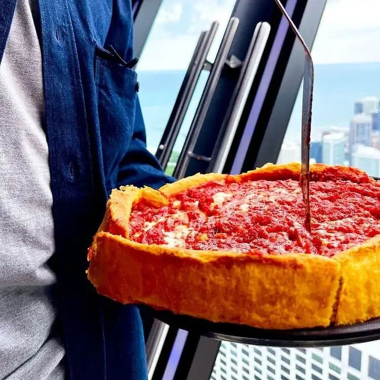 Person holding a large slice of deep-dish pepperoni pizza with a cup of soda on the edge of a building in a city skyline