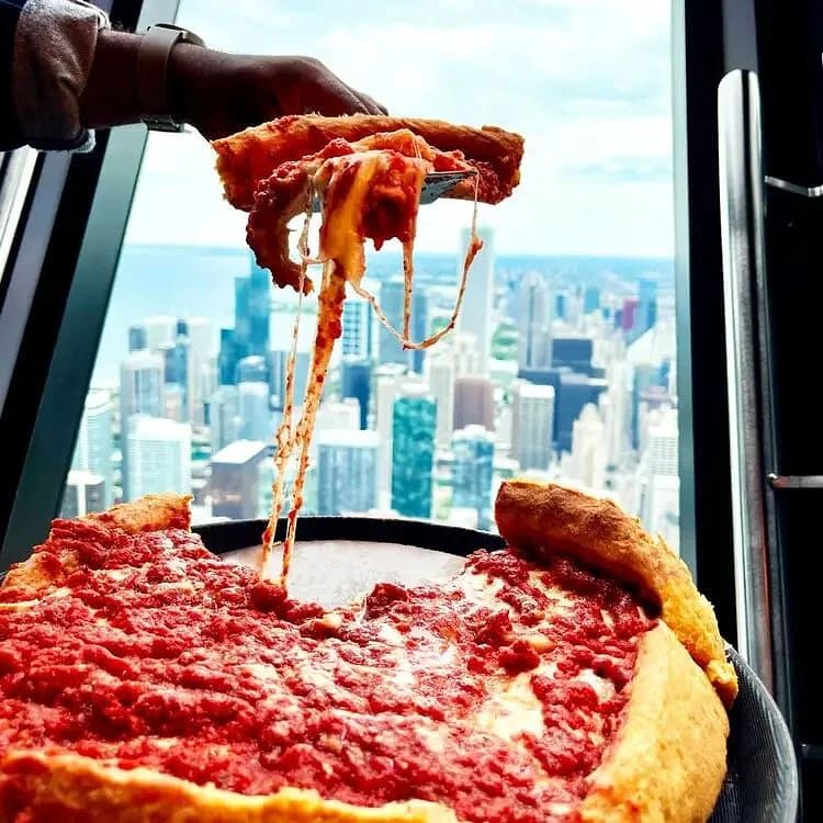 A slice being lifted from a large pepperoni pizza in front of a city skyline view from a tall window.
