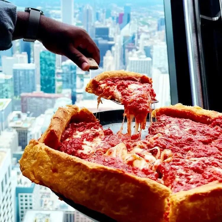 A slice of Chicago-style deep-dish pizza being lifted from a pizza with a cityscape view in the background.