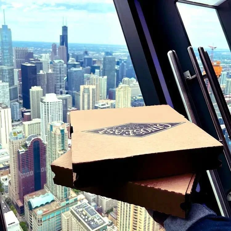 A cardboard pizza box held by a person wearing a glove, with a city skyline visible through a large window in the background.