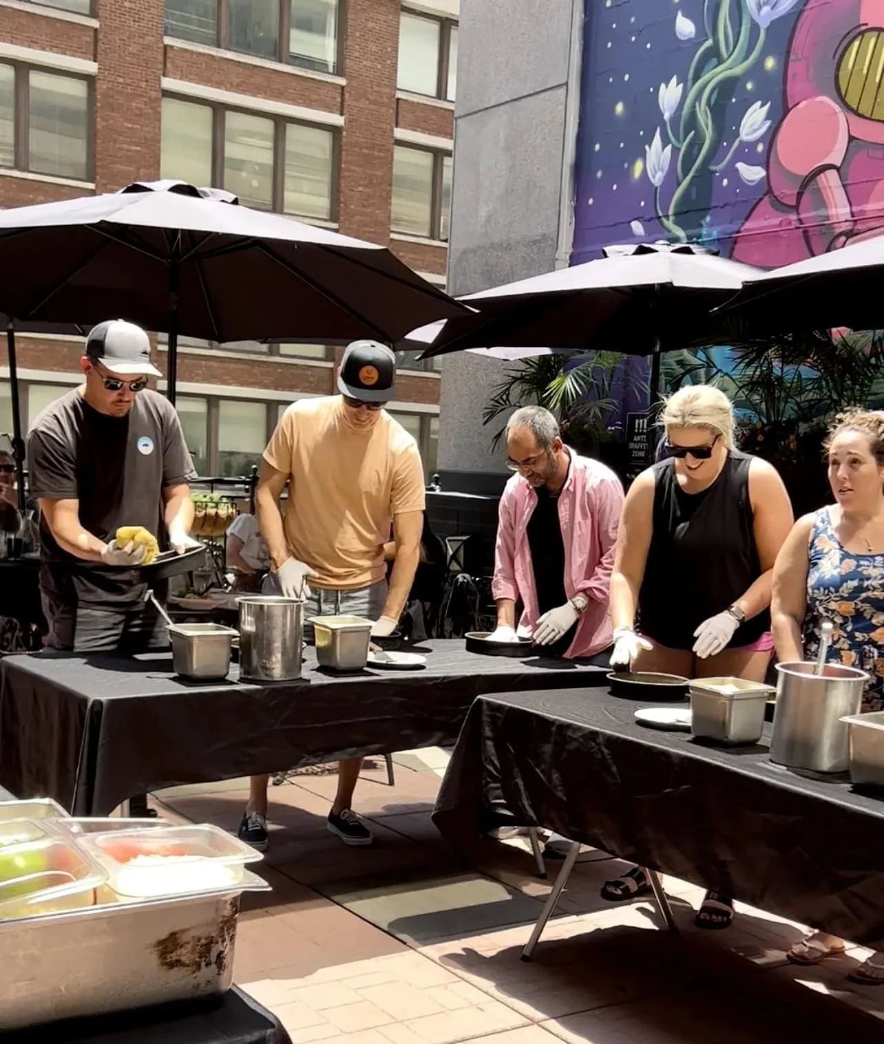 People preparing food at outdoor buffet table under umbrellas, with food containers and plates, in an urban setting.