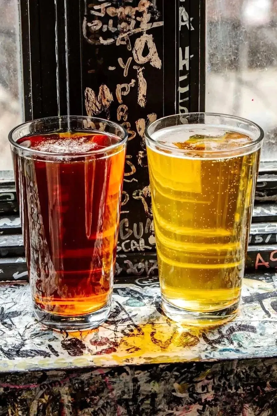 A dark red ale and a light golden beer with a citrus garnish sitting on a graffiti-covered windowsill