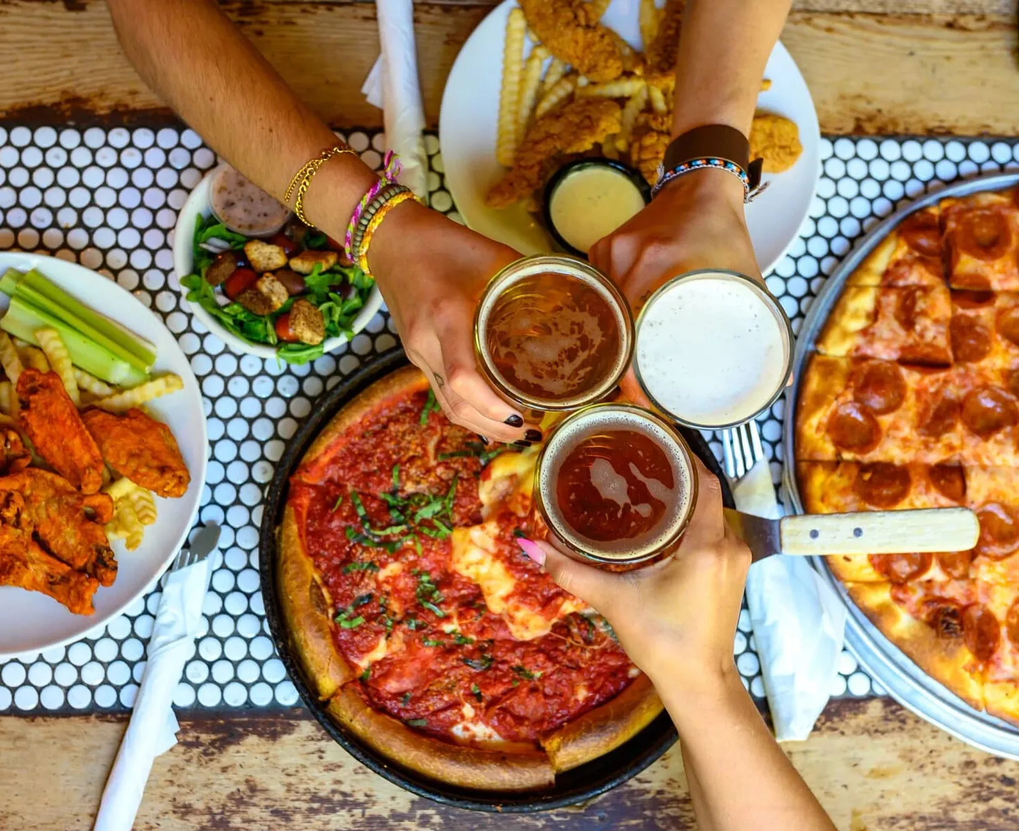 Overhead view of friends clinking glasses of beer over a deep dish pizza in its pan, a pepperoni tavern-style thin crust, wings, a salad and chicken tenders