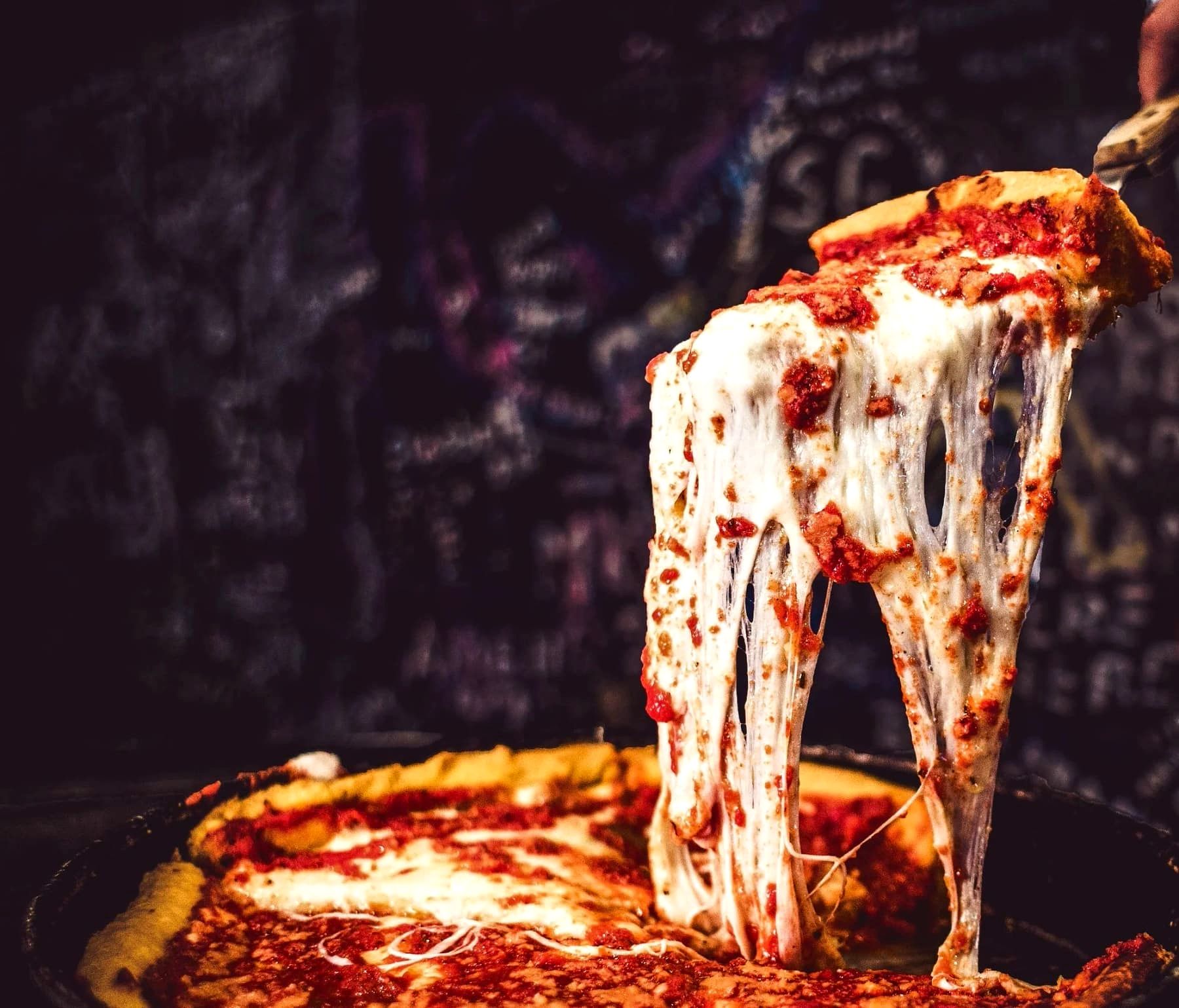 A slice of Gino's East deep dish pizza lifted from the pie with a long, dramatic cheese pull against a graffiti-covered wall