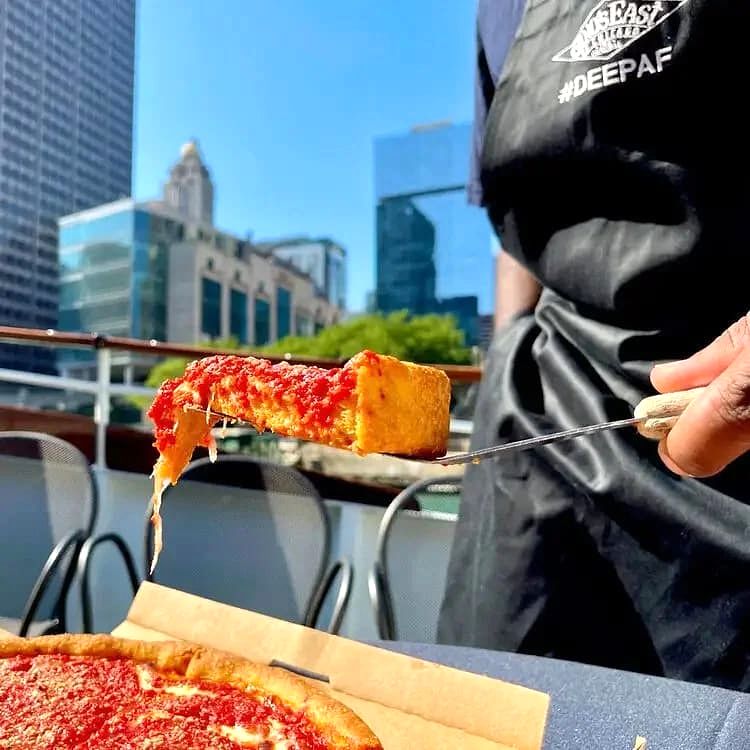 A slice of cheese pizza held up on a metal pizza cutter with city buildings and blue sky behind.