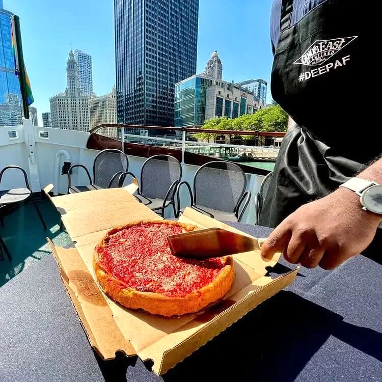 A person slicing a large deep-dish pepperoni pizza on a table outdoors on a boat with city skyscrapers in the background.