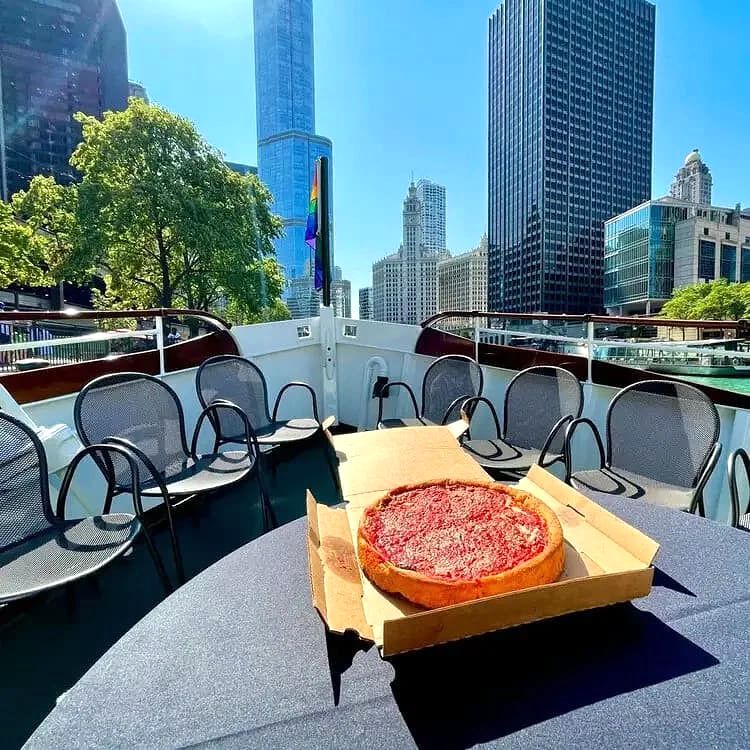 A pizza with red sauce on a table on a boat deck with city buildings and blue sky in the background.