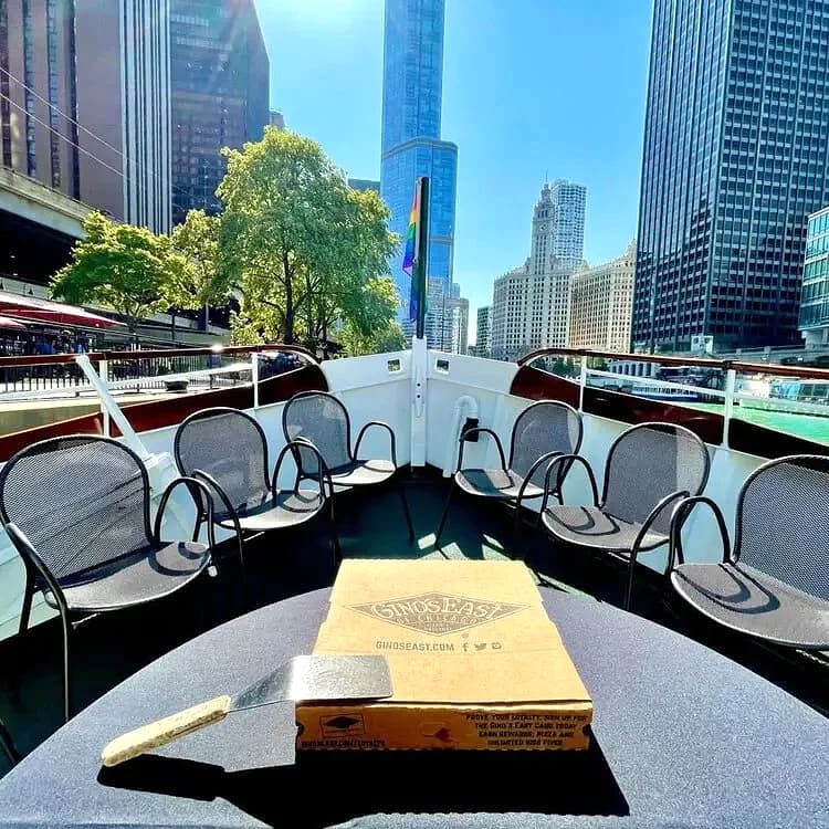 Empty outdoor seating on a boat deck with a pizza box on the table and the Chicago skyline behind.