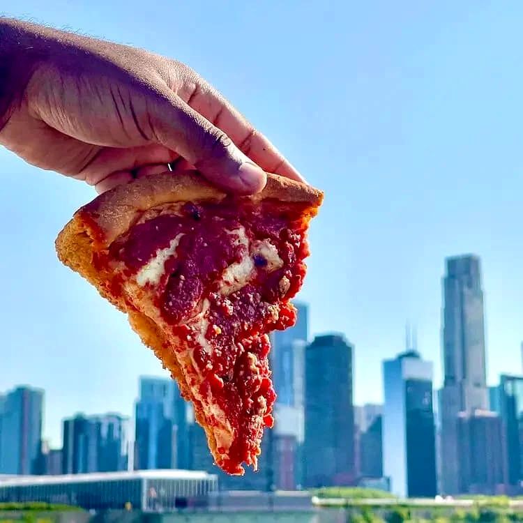 A hand holding a slice of pepperoni pizza against a city skyline with tall buildings and a clear blue sky.
