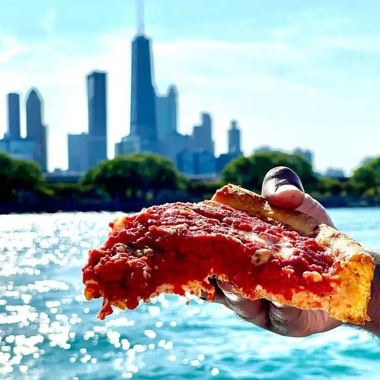A hand holding a slice of pepperoni pizza with a city skyline and water in the background.
