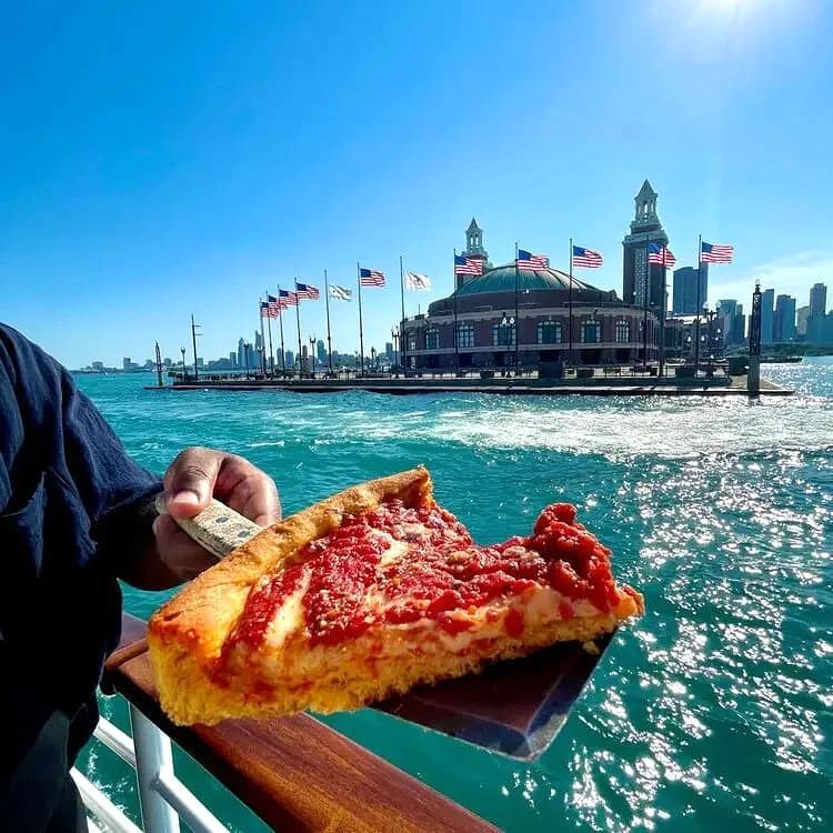Person holding a slice of pepperoni pizza on a boat with a waterfront building and American flags in the background.