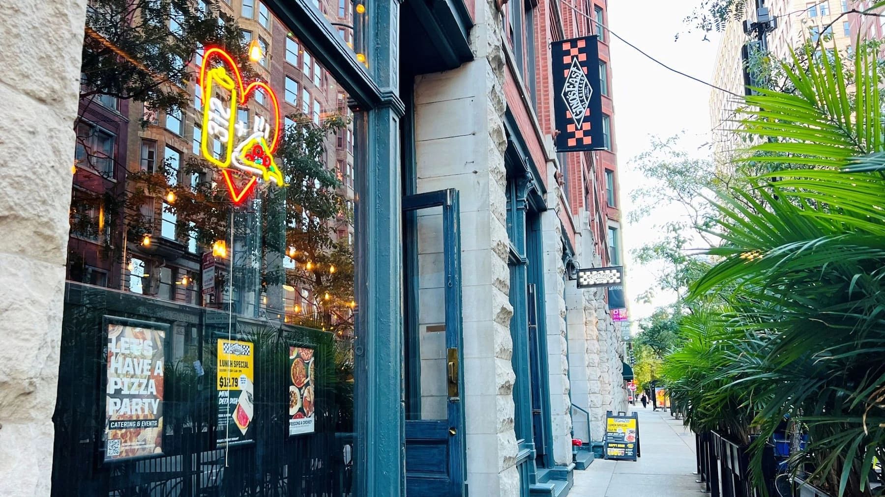Sidewalk view of Gino's East South Loop with palm plantings and a neon pizza sign glowing in the window