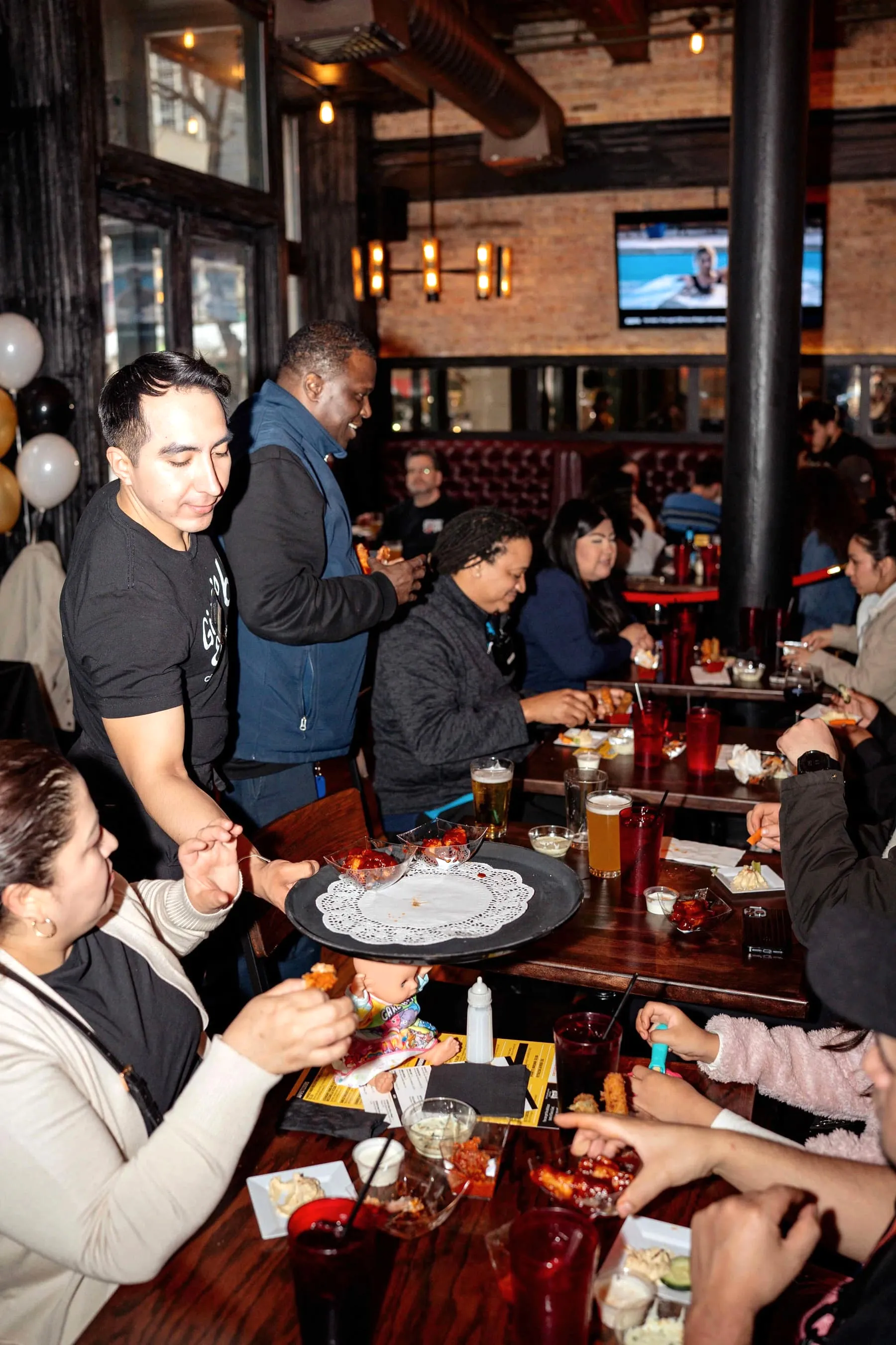 Server carrying a tray of appetizers to a lively group party at Gino’s East South Loop, with balloons, pints of beer and sports on TV